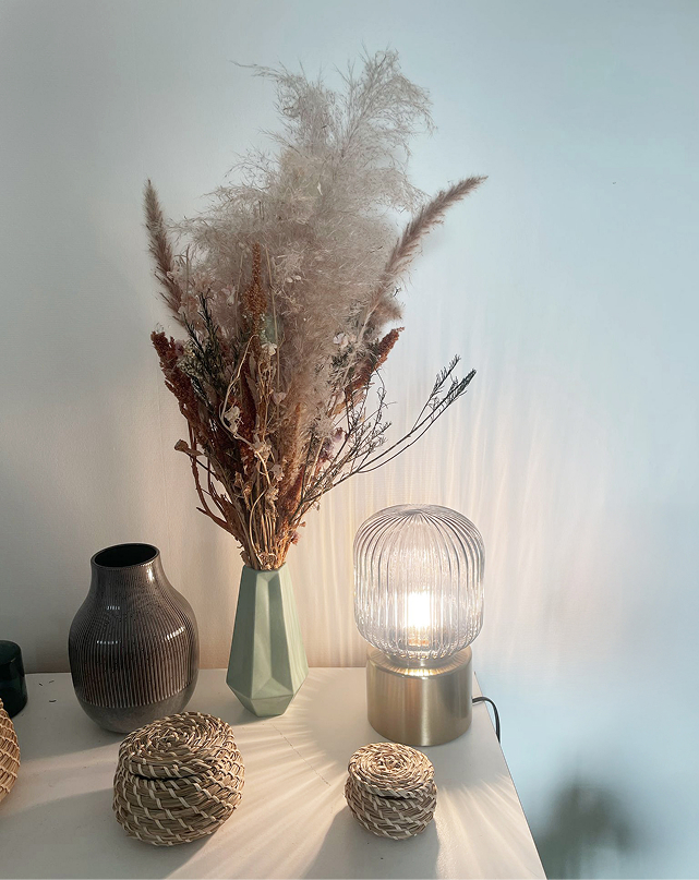 A soft light illuminates decorations on a table. We see a pot of dried flowers, as well as a case and small wicker bowls.