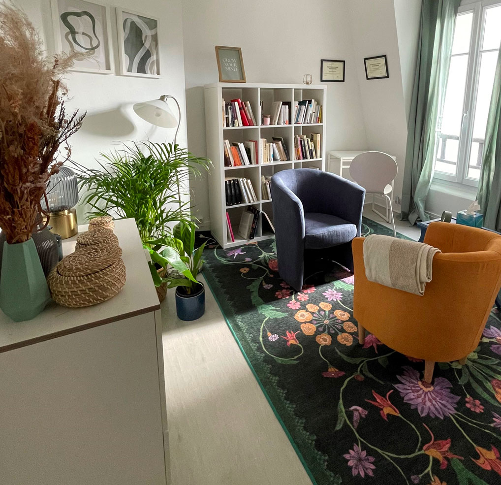 The consulting room: a comfortable armchair in front of a small bookcase, a floral carpet and plants brighten up the room.